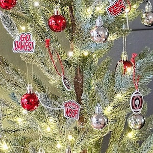 May include: A small artificial Christmas tree decorated with red and silver ornaments, a red and black buffalo plaid bow, and a variety of festive ornaments. The tree is in a burlap pot and is sitting on a wooden table with a black and white beaded garland and a gold and black plaid sign that says "Let's Wassail".