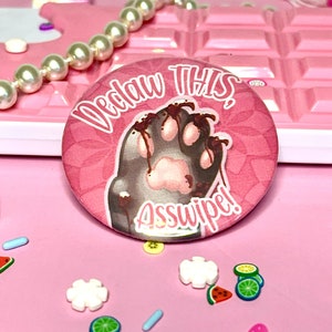 Può includere: Un bottone rosa con un'impronta di zampa di cartone animato con artigli sanguinanti e il testo "Declaw This Asswipe!"