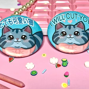 Può includere: Due bottoni rotondi blu e bianchi con un gatto grigio dei cartoni animati che indossa un fiocco rosa. Un bottone dice "I WILL CUT YOU" in lettere rosa con due cuori rosa. L'altro bottone dice "とっても可愛い猫" in lettere nere.