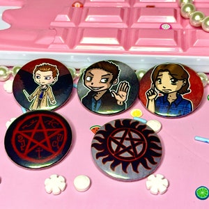 May include: Five round button badges featuring characters and symbols from the TV show Supernatural. The badges are on a pink surface with white sprinkles and candy. The badges feature a pentagram, a symbol with flames, and three characters from the show.