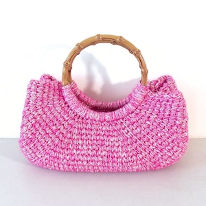 Può includere: Una borsa a mano in crochet rosa con un manico in bambù marrone.