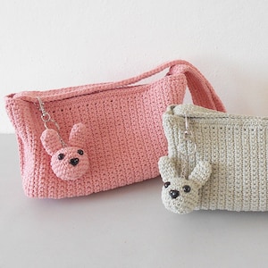 Op de afbeelding: Twee gehaakte handtassen in roze en lichtgroen. Elke tas heeft een rechthoekige vorm met een ritssluiting en een bijpassende band. Een kleine konijnenhanger hangt aan de rits. De tassen zijn handgemaakt en hebben een getextureerde uitstraling.