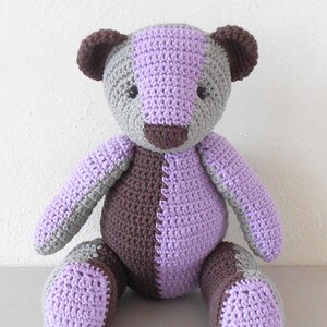 Op de afbeelding: Een handgemaakte gehaakte teddybeer in tinten grijs, bruin en lavendel. De beer heeft een tweekleurig ontwerp, met één kant bruin en lavendel, en de andere kant grijs en lavendel. De beer zit rechtop.