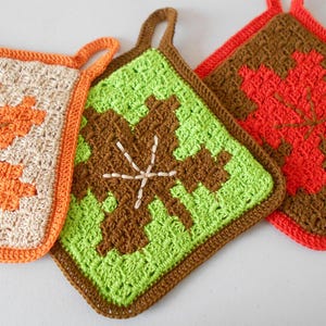 May include: Three handmade crochet potholders with leaf designs. One is beige with an orange leaf, another is green with a brown leaf, and the third is brown with an orange leaf. Each has a loop for hanging.