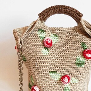 Puede incluir: Un bolso de crochet con un diseño floral con flores rojas, blancas y verdes. El bolso tiene un asa marrón y una correa de cadena dorada.