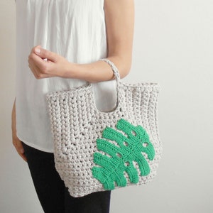 May include: A crocheted tote bag with a green leaf design. The bag is made of a thick, textured yarn and has a simple, casual style.