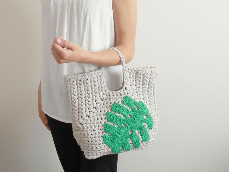May include: A crocheted tote bag with a green leaf design. The bag is made of a thick, textured yarn and has a simple, casual style.