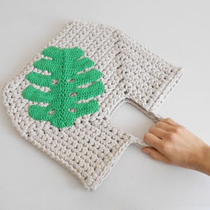 May include: A crocheted tote bag with a green leaf design. The bag has a single handle and is made of a natural beige yarn.