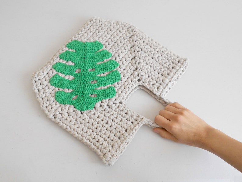 May include: A crocheted tote bag with a green leaf design. The bag has a single handle and is made of a natural beige yarn.
