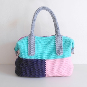 Puede incluir: Bolso tote de ganchillo hecho a mano con un diseño de bloques de color. La sección superior es turquesa, la del medio es gris claro y la inferior está dividida en secciones azul marino y rosa. El bolso tiene dos asas grises y un cierre de cremallera.