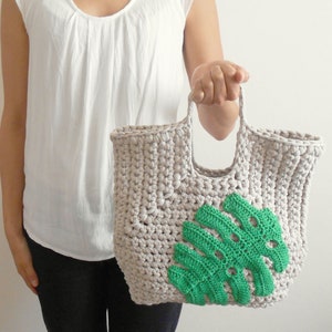 May include: A crocheted tote bag with a green monstera leaf design. The bag is made of beige yarn and has a wide, flat handle.