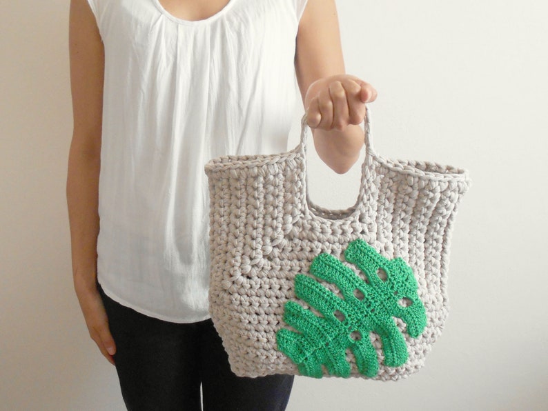 May include: A crocheted tote bag with a green monstera leaf design. The bag is made of beige yarn and has a wide, flat handle.