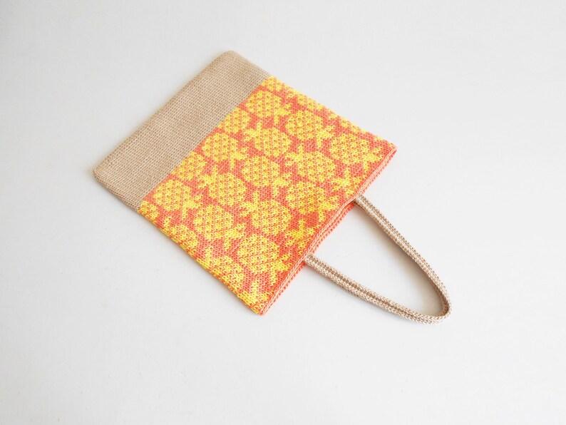 May include: A crocheted tote bag with a yellow and orange pineapple pattern. The bag has a natural jute handle.