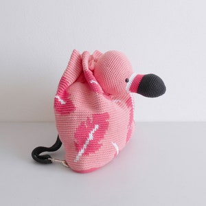 以下が含まれることがあります： 黒いストラップが付いたピンクのクロシェ編みフラミンゴのバックパック。バックパックは、ピンクの体と黒いクチバシを持つフラミンゴの形をしています。