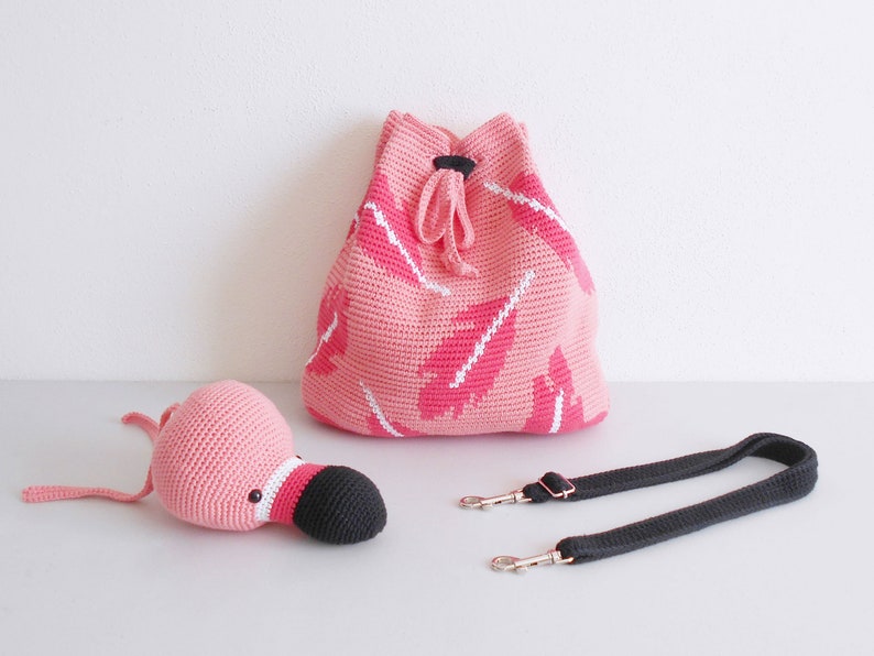 以下が含まれることがあります： 黒いクチバシのピンクの編みぐるみフラミンゴと、黒いストラップのついたピンクの編みぐるみ巾着袋。袋にはピンクと白の羽根模様があります。
