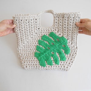 May include: A crocheted tote bag with a green monstera leaf design. The bag is made of cream-colored yarn and has a single handle.