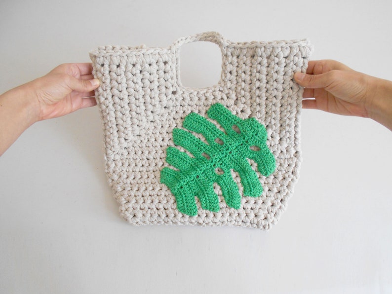 May include: A crocheted tote bag with a green monstera leaf design. The bag is made of cream-colored yarn and has a single handle.