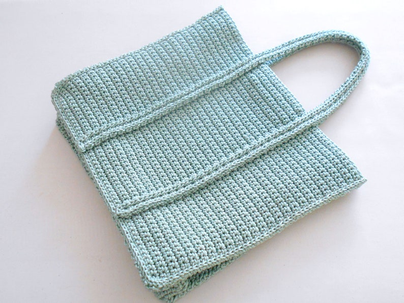 Op de afbeelding: Een lichtblauwe gehaakte tote bag met twee handvatten. De tas is gemaakt met een eenvoudig steekpatroon en heeft een rechthoekige vorm.