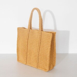 Op de afbeelding: Een gehaakte tote bag in een lichtgele kleur. De tas heeft een brede, platte basis en een rechthoekige vorm. De tas heeft een enkele handvat die aan de bovenkant van de tas is bevestigd.