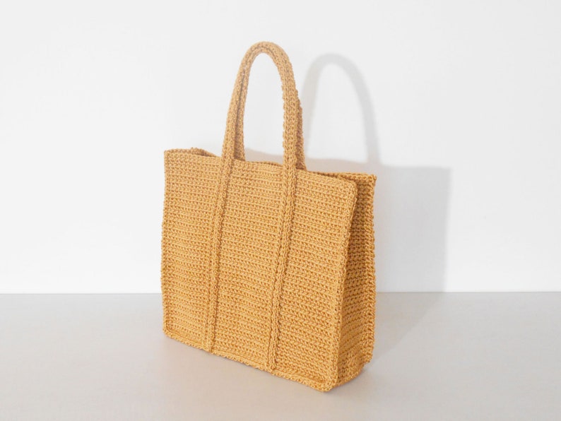 Op de afbeelding: Een gehaakte tote bag in een lichtgele kleur. De tas heeft een brede, platte basis en een rechthoekige vorm. De tas heeft een enkele handvat die aan de bovenkant van de tas is bevestigd.