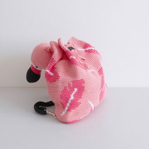 以下が含まれることがあります： 白い葉のデザインが施されたピンクのフラミンゴの形をしたかぎ針編みのバックパック。バックパックには黒いストラップが付いています。