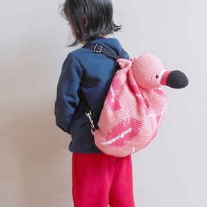 以下が含まれることがあります： 黒い目と黒いクチバシが付いたピンクの編みぐるみフラミンゴのバックパック。バックパックは子供が背負っています。