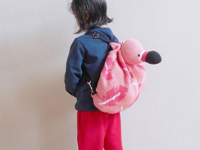 以下が含まれることがあります： 黒い目と黒いクチバシが付いたピンクの編みぐるみフラミンゴのバックパック。バックパックは子供が背負っています。