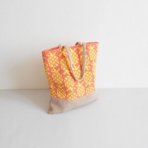 May include: A crocheted tote bag with a yellow and orange geometric pattern. The bag has a beige bottom and two beige straps.