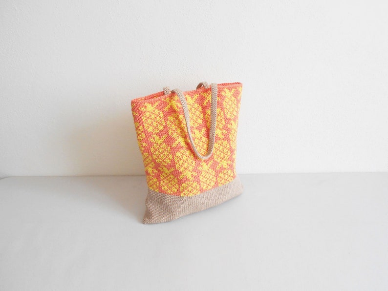 May include: A crocheted tote bag with a yellow and orange geometric pattern. The bag has a beige bottom and two beige straps.