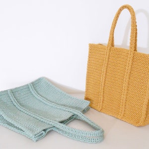 Op de afbeelding: Twee gebreide boodschappentassen, een in lichtblauw en een in mosterdgeel. Beide tassen hebben een eenvoudig ontwerp met verticale strepen.