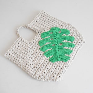 May include: A crocheted tote bag with a green leaf design. The bag is made of natural cotton yarn and has a single handle.