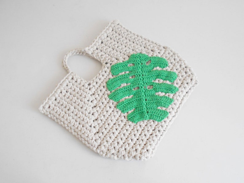 May include: A crocheted tote bag with a green leaf design. The bag is made of natural cotton yarn and has a single handle.