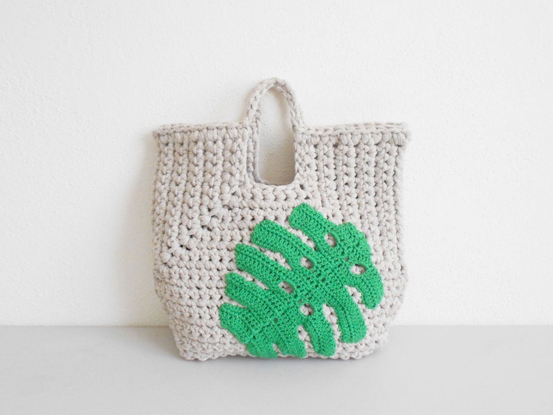 May include: A crocheted tote bag with a green leaf design. The bag is made of beige yarn and has a single handle.