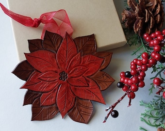 May include: A handcrafted, red and brown poinsettia ornament with detailed leaf patterns. A beige gift box tied with gold string and a red ribbon sits nearby. Festive greenery, pinecones, and red berries complete the holiday arrangement.