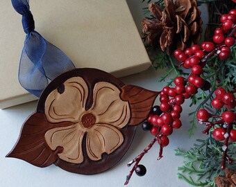 May include: A brown leather ornament shaped like a dogwood flower with a light brown center. The ornament is on a white surface with a blue ribbon and a brown gift box.