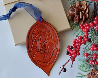 May include: A brown leather Christmas ornament shaped like a teardrop with a carved image of Mary and baby Jesus. The ornament is hanging from a blue ribbon and is surrounded by a Christmas tree branch with red berries.