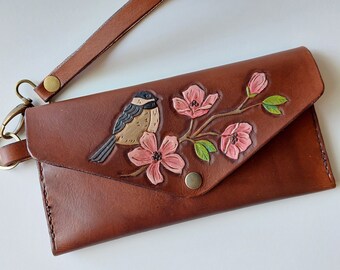 May include: A brown leather wallet with a floral design and a small bird. The wallet has a flap closure and a wrist strap.
