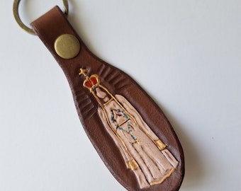 May include: Brown leather keychain with a gold-painted image of Our Lady of Fatima, a religious figure wearing a white robe and a crown.