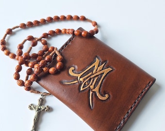 May include: A brown leather rosary case with an embossed gold design and a wooden bead rosary. A metal crucifix hangs from the rosary. The case and rosary are set against a white background.