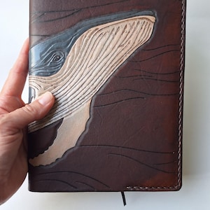 May include: A brown leather journal with a detailed carving of a whale's head. The whale is depicted in shades of gray and beige, with the surrounding leather textured to resemble water. The journal has a stitched border and a black ribbon bookmark.