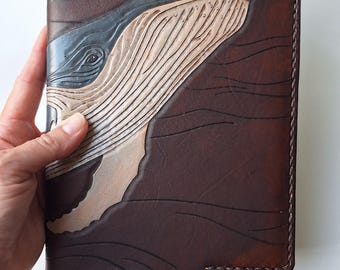 May include: A brown leather journal with a detailed carving of a whale's head. The whale is depicted in shades of gray and beige, with the surrounding leather textured to resemble water. The journal has a stitched border and a black ribbon bookmark.