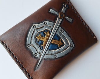 May include: A brown leather wallet with a tooled design of a silver shield and sword with a gold cross.