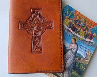 May include: A handcrafted, brown leather journal cover with a Celtic cross design embossed on the front. The cover is next to a magazine with the title "Magnificat" and the date "December 2022". The journal cover is a religious item.