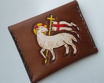 May include: A brown leather wallet with a tooled design of a white lamb with a halo and a red and white flag. The lamb is standing on a yellow cross.