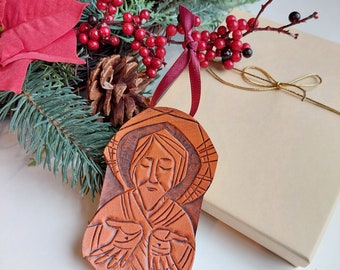 May include: A handcrafted, brown leather Christmas ornament featuring a carved image of a religious figure with outstretched hands. The ornament is suspended by a burgundy ribbon and is surrounded by festive greenery, red berries, and a gift box.
