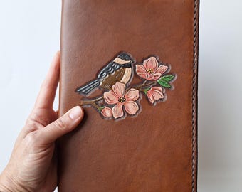 May include: A brown leather journal cover with a hand-tooled design of a bird perched on a flowering branch. The bird is brown, black, and white, and the flowers are pink. The cover is rectangular and has stitched edges.