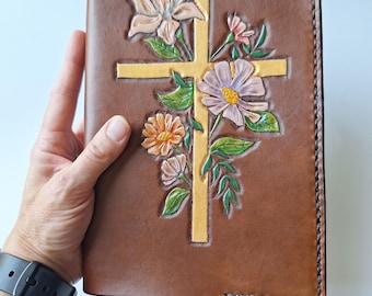 May include: A brown leather book cover featuring a hand-tooled design of a gold-colored cross intertwined with various flowers in shades of pink, orange, and green. The cover has visible dark stitching along the right edge and a smooth finish.