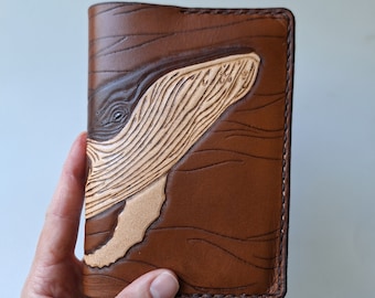 May include: A brown leather journal with a detailed whale design. The whale is carved in a lighter tan color, with the body and background in a darker brown. The journal is held in a hand.