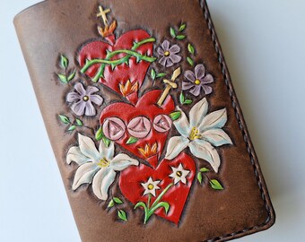May include: A brown leather journal cover with a tooled design of three red hearts with thorns, a cross, and white and purple flowers.