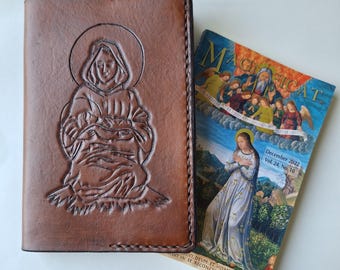 May include: A brown leather journal cover with an embossed image of a religious figure. Next to it is a magazine titled "Magnificat" with a religious illustration and the text "December 2022 Vol. 24, No. 10". The journal cover has stitched edges.